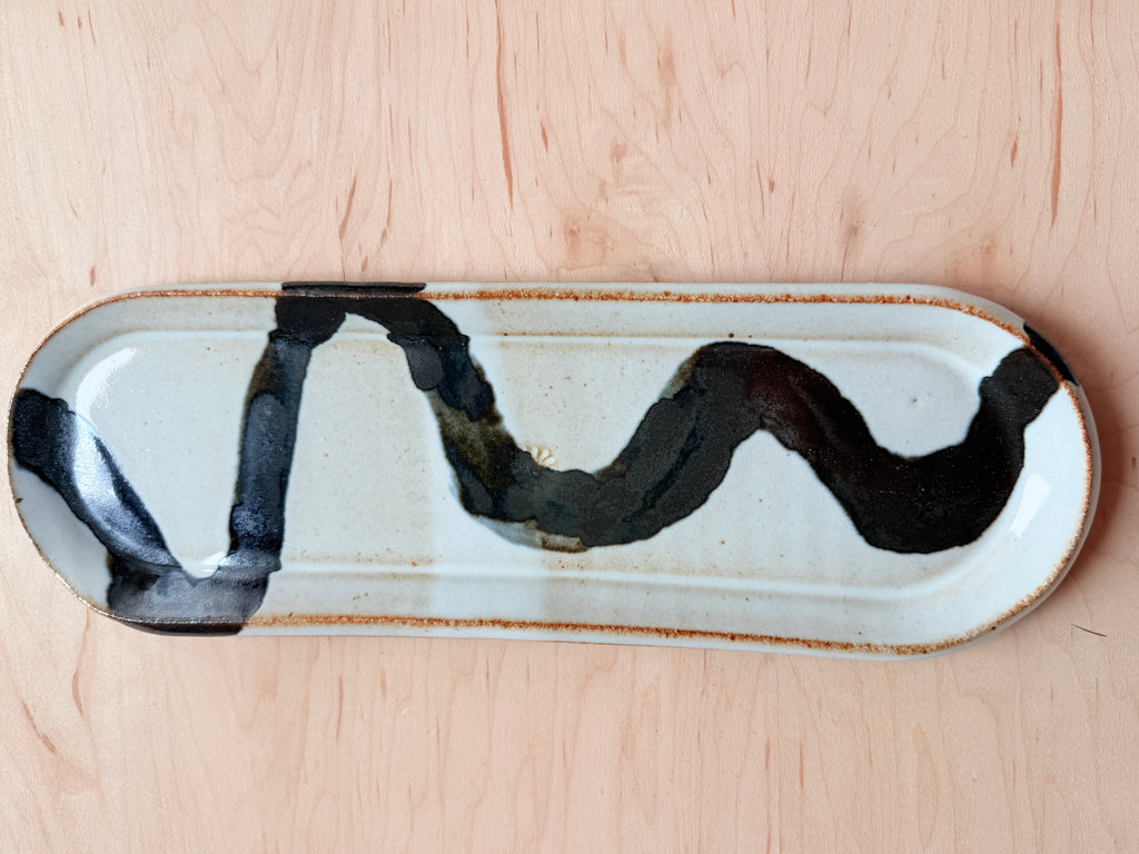 A rectangular ceramic dish with rounded corners, featuring a glossy white glaze, a wavy black brushstroke design, and a rustic brown rim, shown on a light wood background.