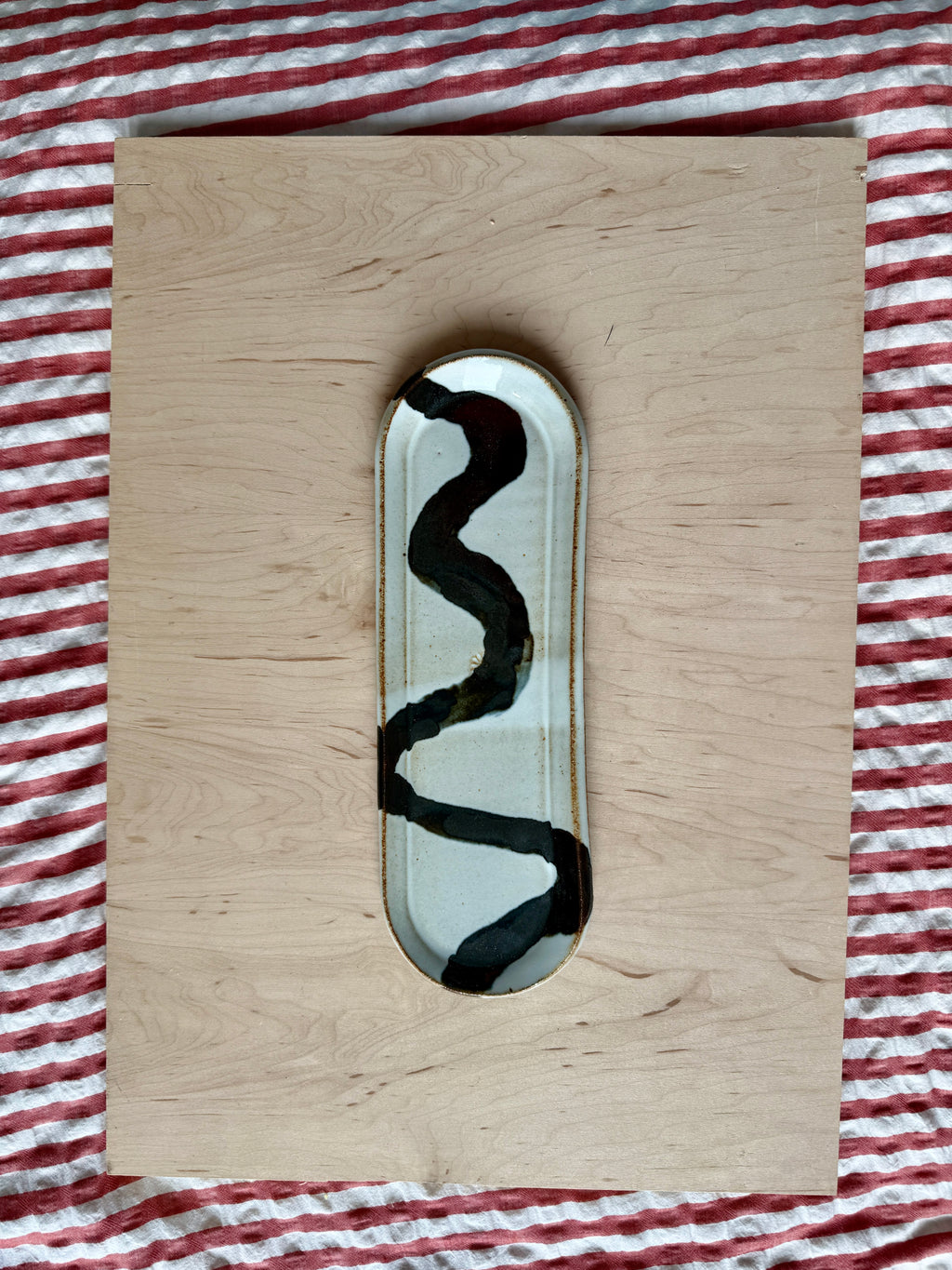 A rectangular light wood board with a small oval inset featuring a black wavy line pattern on a white background, placed on a red and white striped fabric surface.