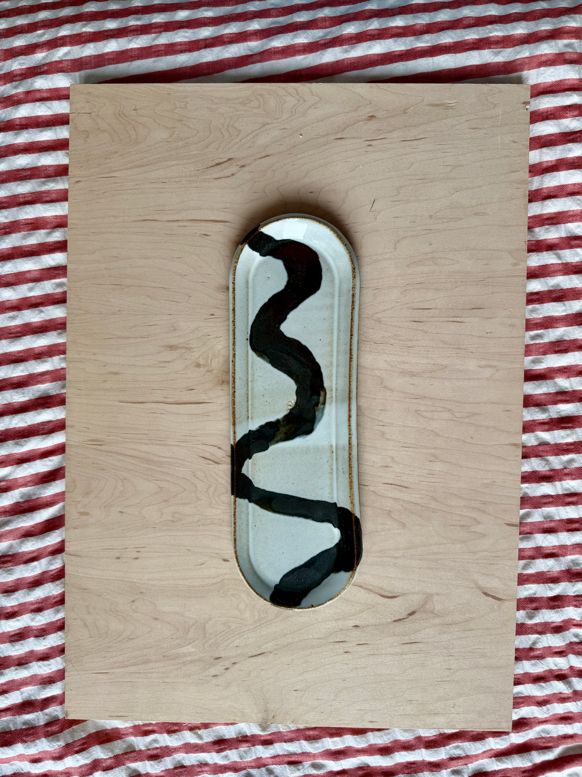 A rectangular light wood board with a small oval inset featuring a black wavy line pattern on a white background, placed on a red and white striped fabric surface.