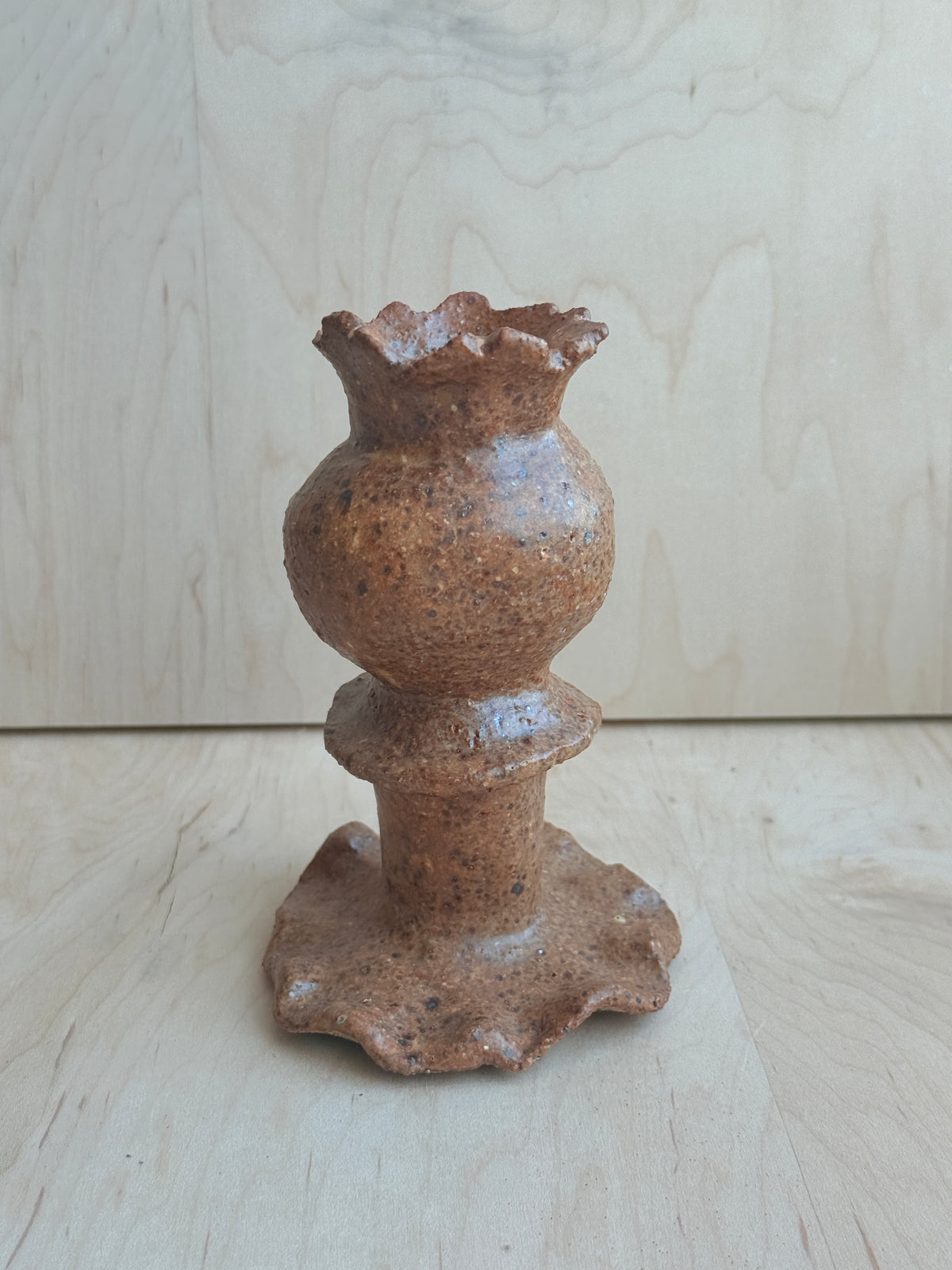 A handmade ceramic candle holder with a textured, speckled brown glaze, featuring a bulbous body and a flared, scalloped base, set against a light wood grain background.