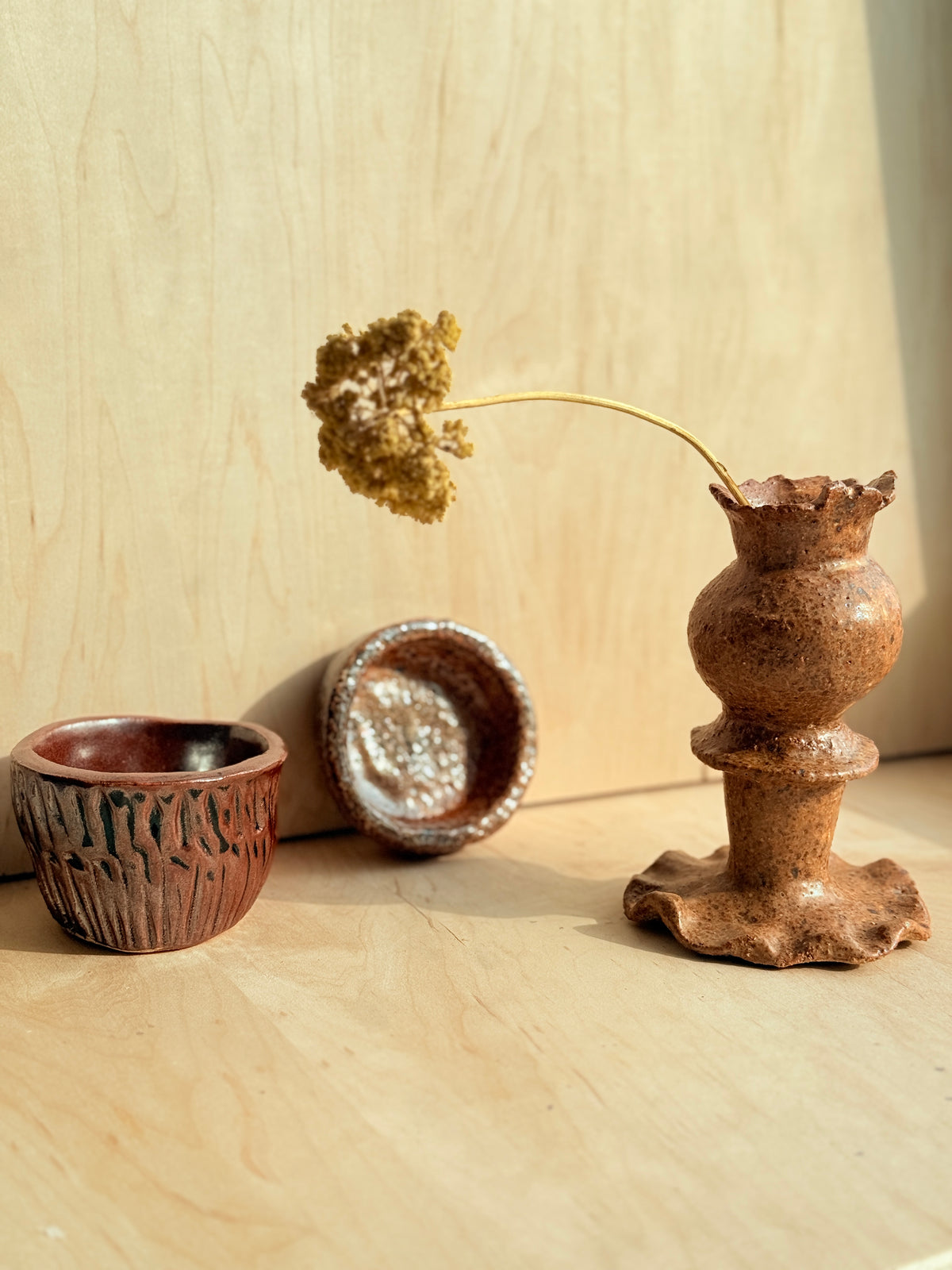 Three handmade stoneware vessels in earthy terracotta and brown glazes arranged on a light wood surface against a wood-paneled background. One tall pedestal-style vase holds a dried yellow-green stem, with two smaller bowls nearby.