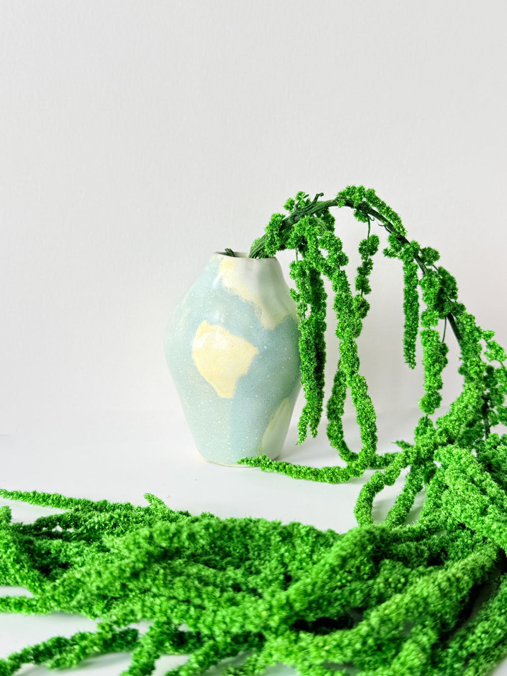 Small handmade bud vase in blue and butter yellow with green dried amaranth draped across a white surface