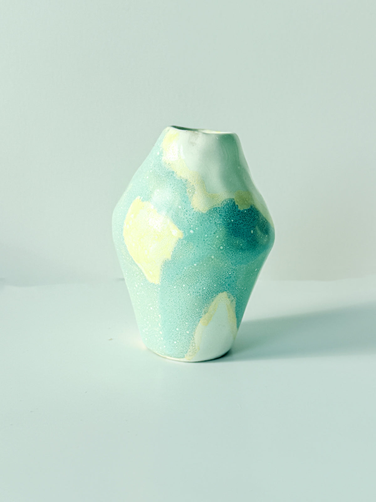 Front view of a small handmade ceramic bud vase with butter yellow glaze patches on a blue speckled body