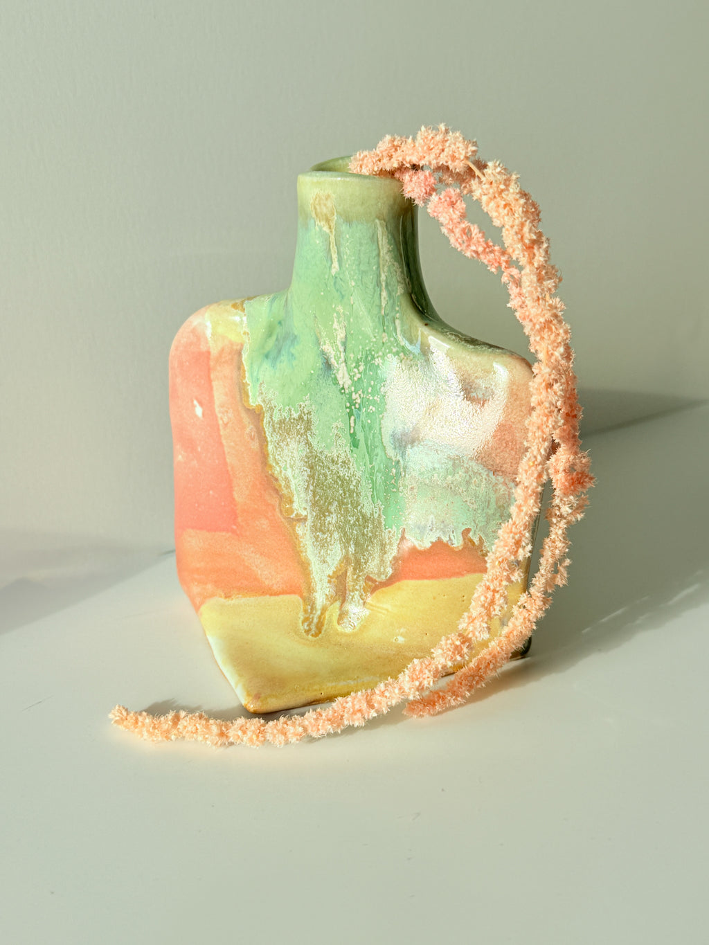 A small sculptural ceramic vase with an abstract multicolor glaze in green, peach, orange, and yellow, decorated with a long textured peach string draped over it, set against a plain light gray background.