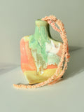 A small sculptural ceramic vase with an abstract multicolor glaze in green, peach, orange, and yellow, decorated with a long textured peach string draped over it, set against a plain light gray background.