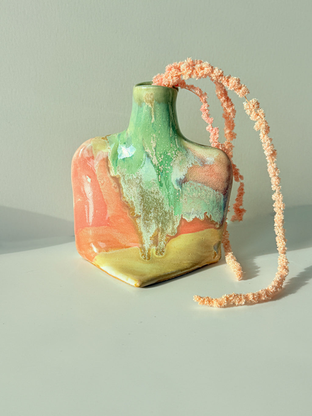A small ceramic bottle-shaped bag with a glossy abstract glaze in green, peach, orange, and gold, featuring a chunky peach woven strap, set against a neutral light gray background.