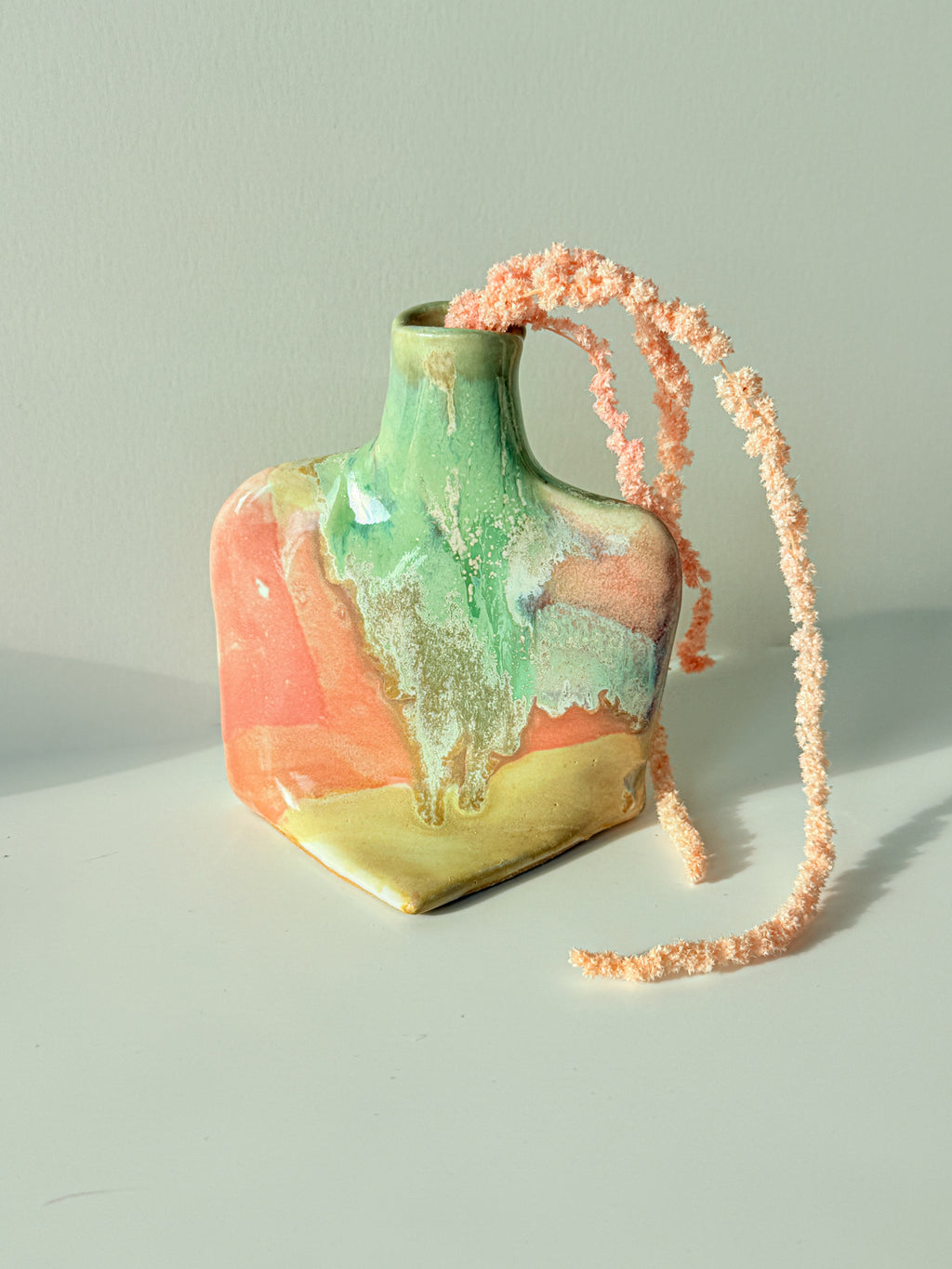 A small ceramic vase with a glossy marbled finish in green, peach, and gold, featuring a long, fuzzy peach-pink braided strap draped over it, set against a plain light gray background.