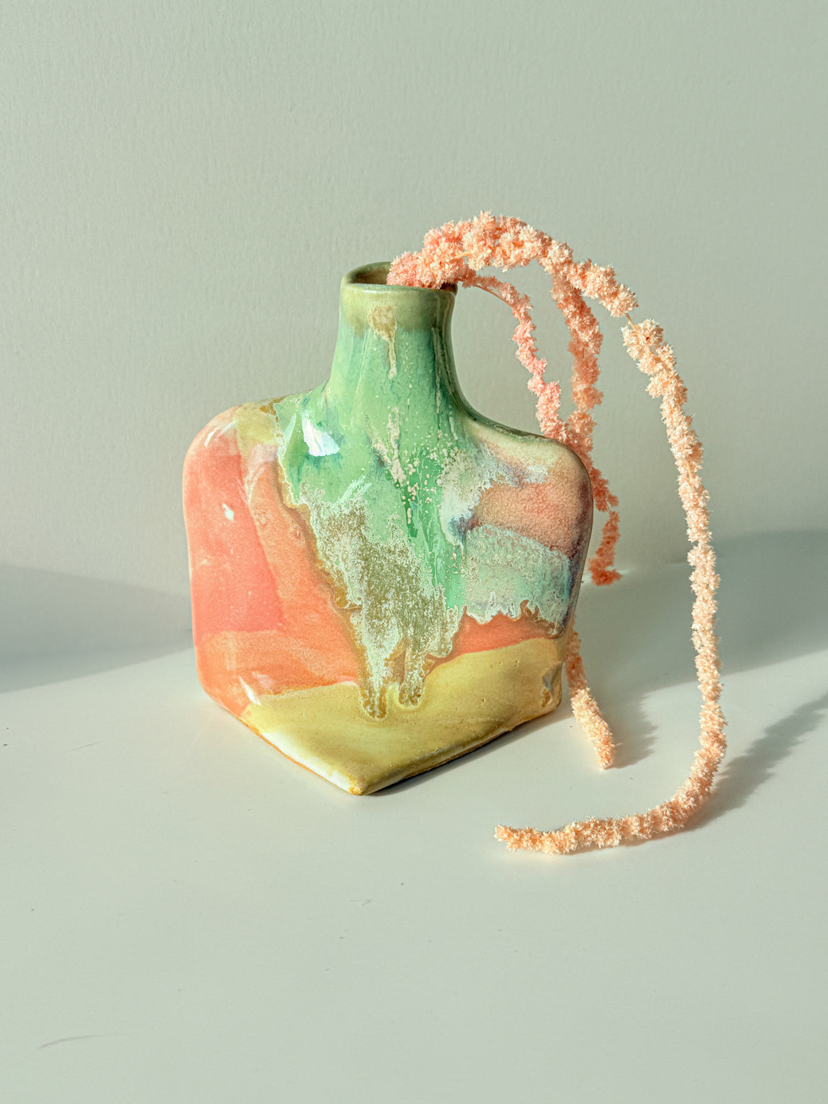 A small ceramic vase with a glossy marbled finish in green, peach, and gold, featuring a long, fuzzy peach-pink braided strap draped over it, set against a plain light gray background.