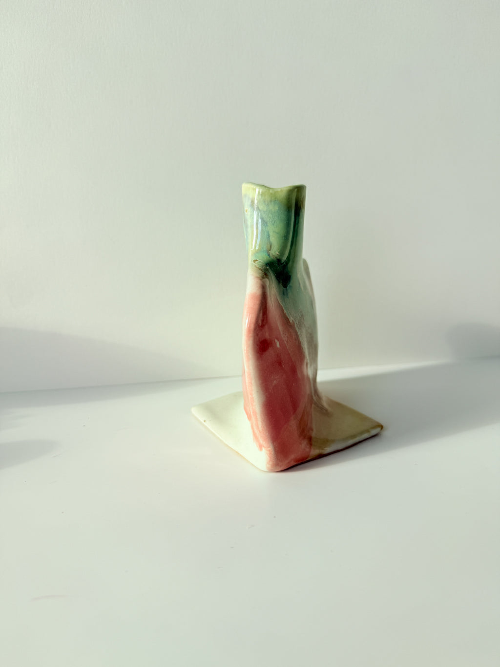 A small sculptural ceramic vase with a square base, featuring a glossy marbled glaze in pink, green, cream, and white, set against a plain white background.