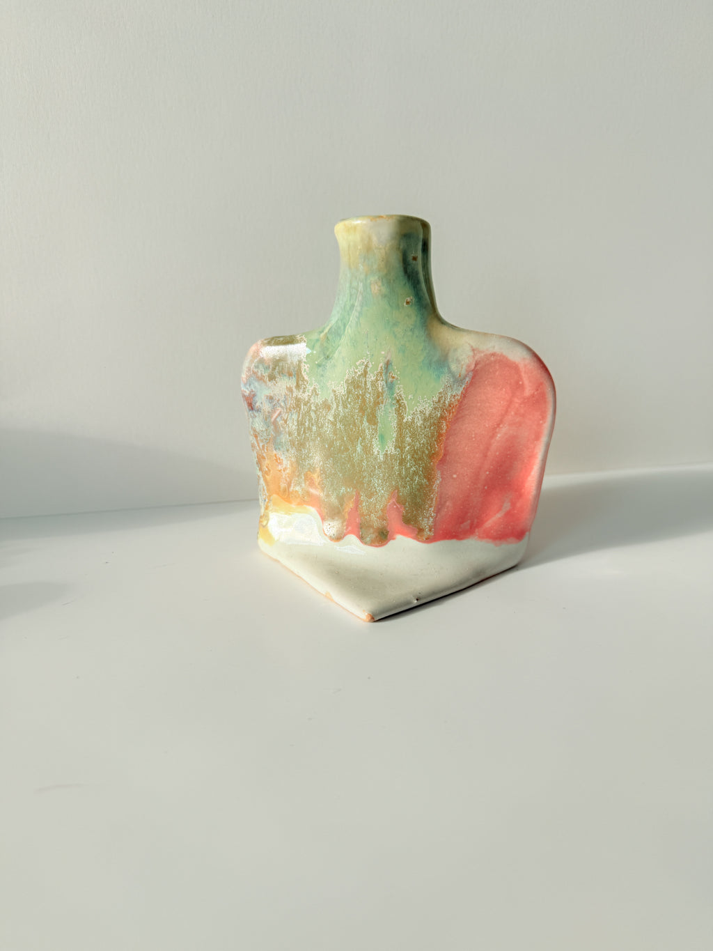 A small handmade ceramic vase with a wide, rounded body and a narrow neck, decorated with a multicolor glaze featuring pink, green, gold, and white.