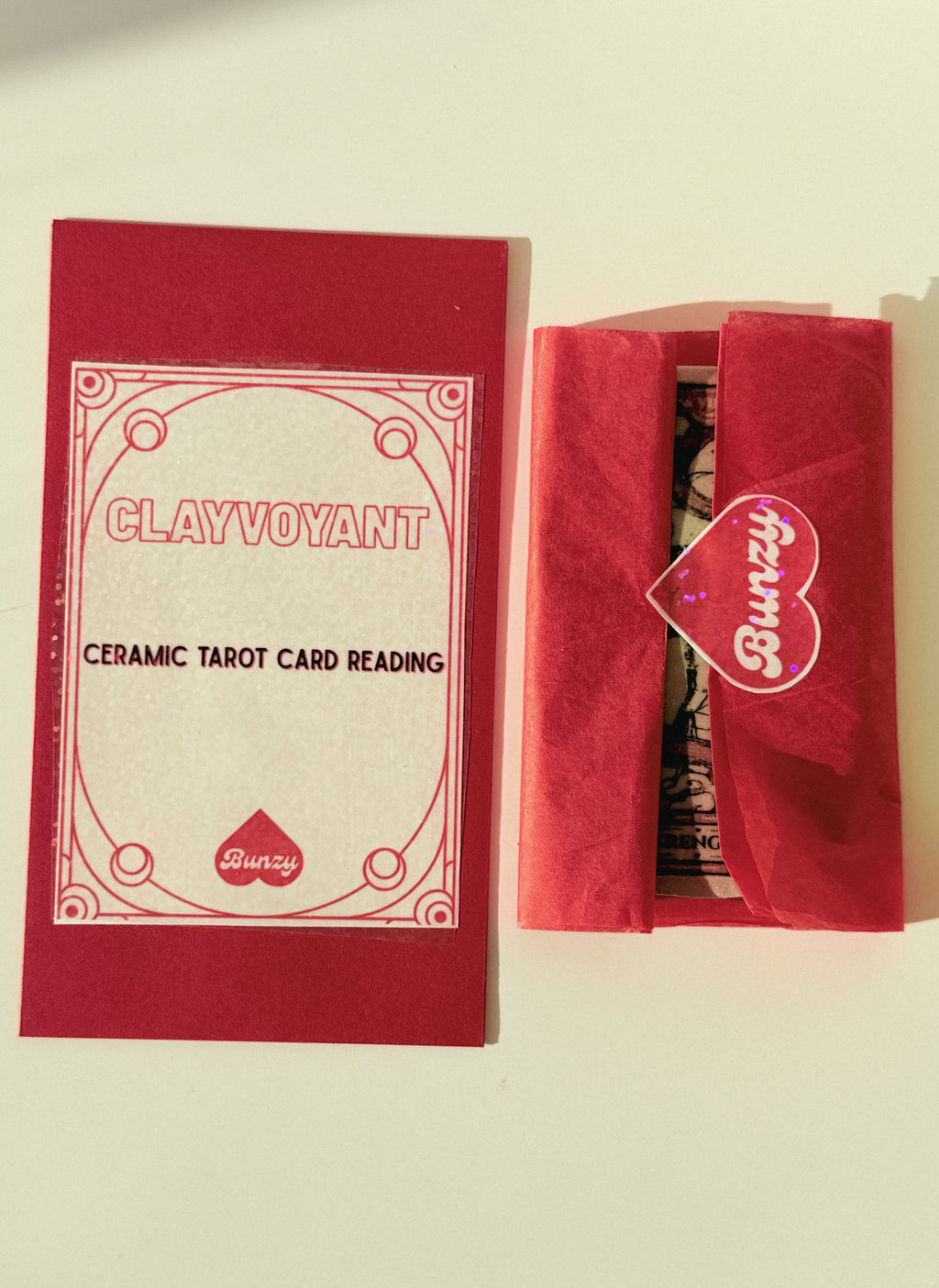 A red and cream tarot card reading kit by Clayvoyant, shown with its outer sleeve and a tissue-wrapped box on a light beige background.