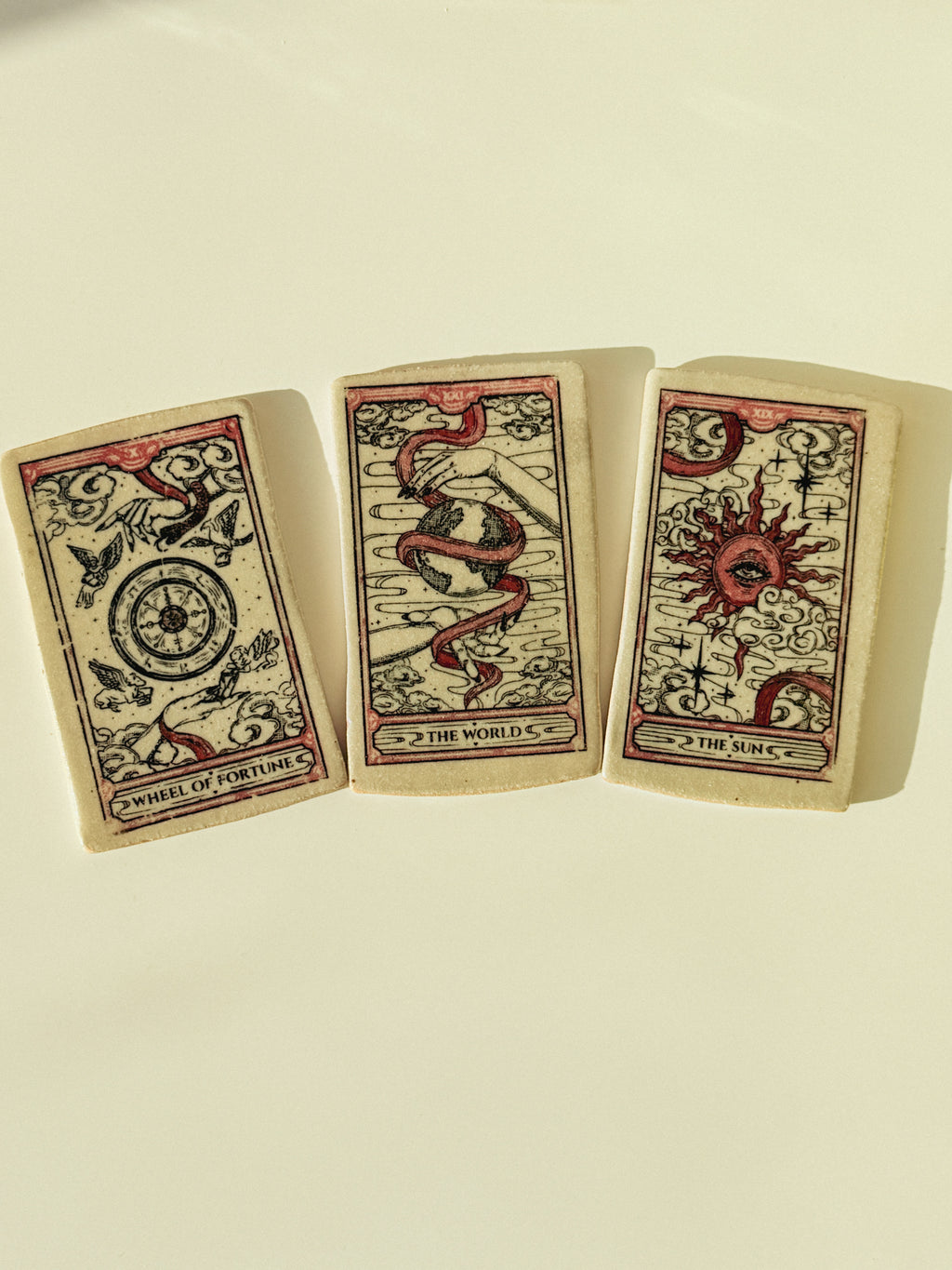 Three illustrated ceramic tarot cards arranged side by side on a plain cream background. The cards show vintage-style artwork with black linework and red accents, including the titles Wheel of Fortune, The World, and The Sun.