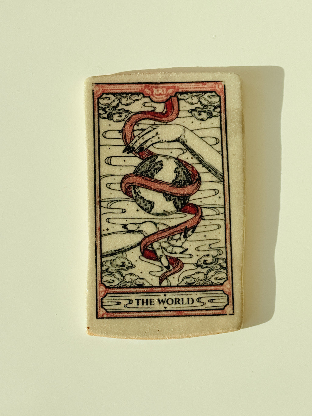 A square ceramic tile featuring the 'The World' tarot card design, with a red snake wrapping around a globe and black line art on a cream background, photographed against a light beige wall with a soft shadow.