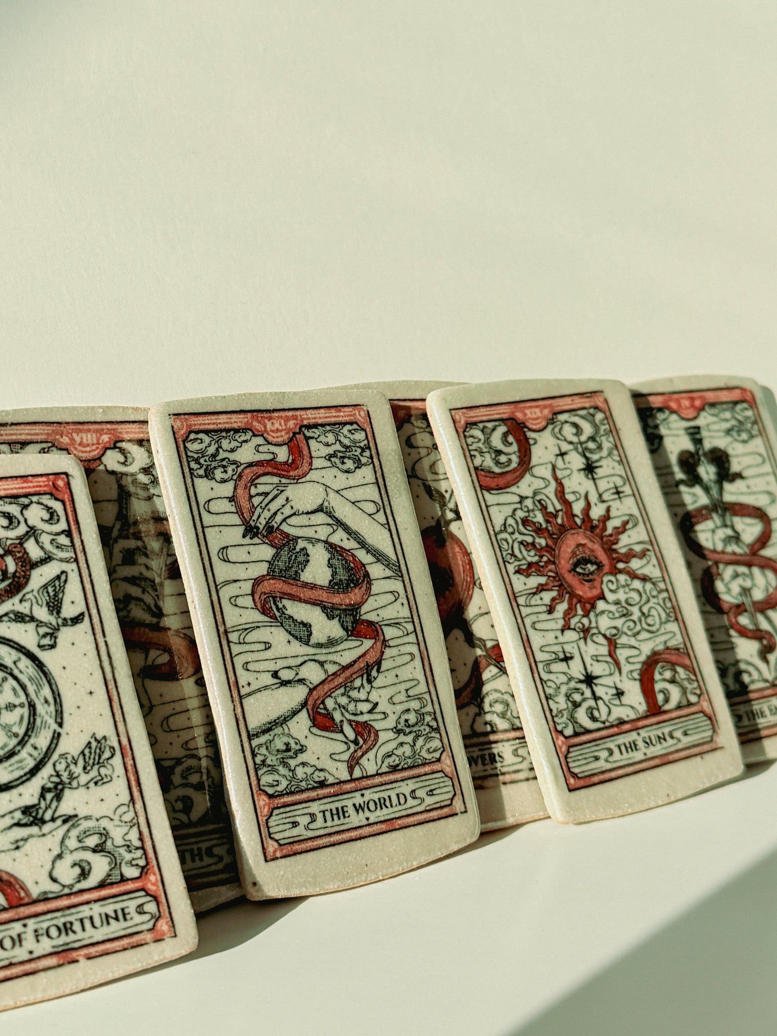 A row of illustrated ceramic tarot cards fanned out on a light surface, showing red and black artwork on aged off-white card stock. Visible card titles include The World, The Sun, and The Hanged Man.