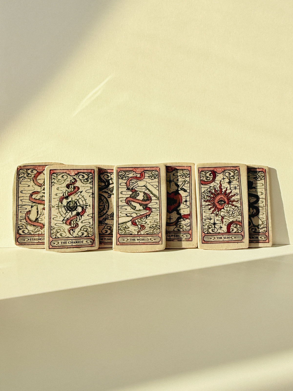 A row of tarot cards fanned out on a warm cream-colored surface, with dramatic diagonal sunlight casting shadows. The cards feature vintage-style illustrations in red, black, and beige, showing symbols like a snake, a sun, and a skull.