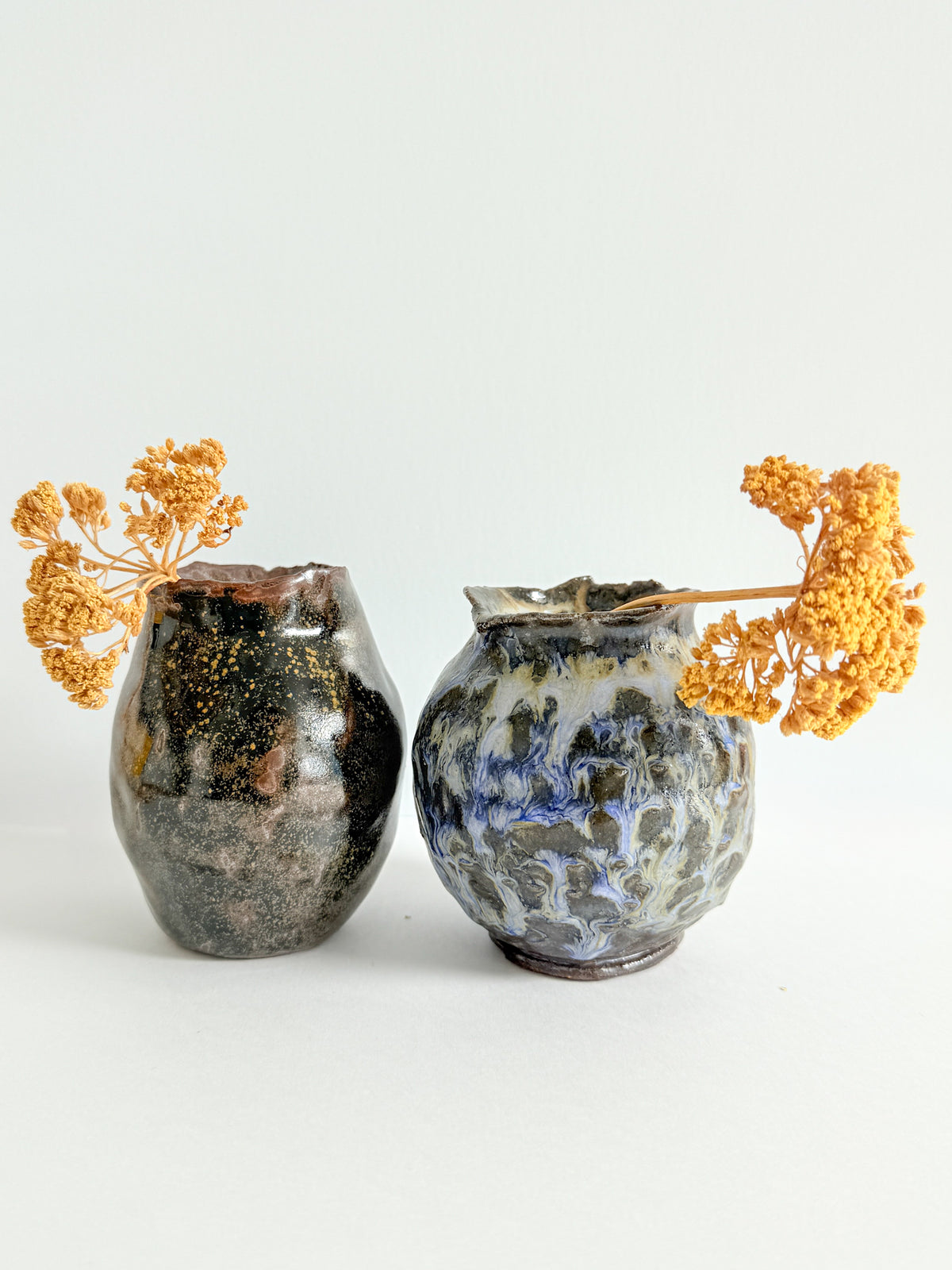 Two handbuilt stoneware bud vases by Bunzy NYC holding dried yellow yarrow, one in glossy speckled brown glaze and one in purple and cream drip glaze.