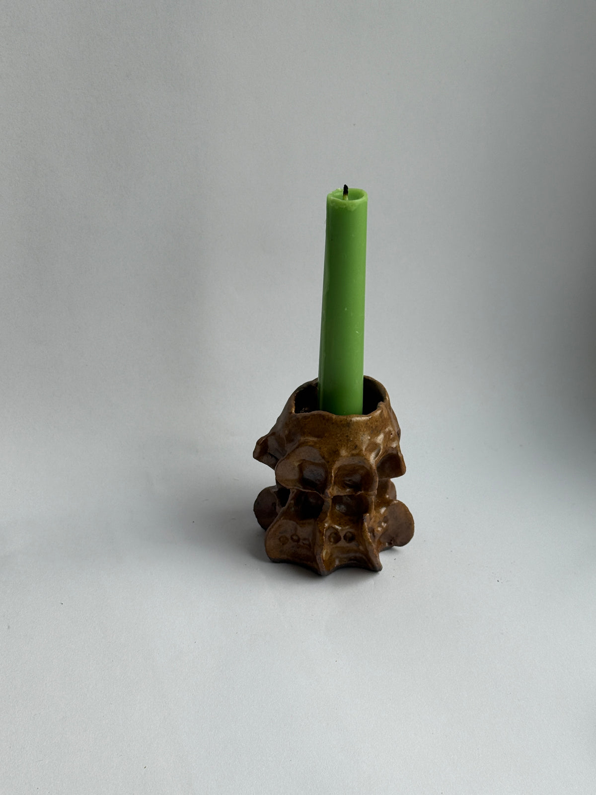 A green taper candle standing upright in a brown, organic-shaped holder with a textured, organic surface, set against a neutral gray background.