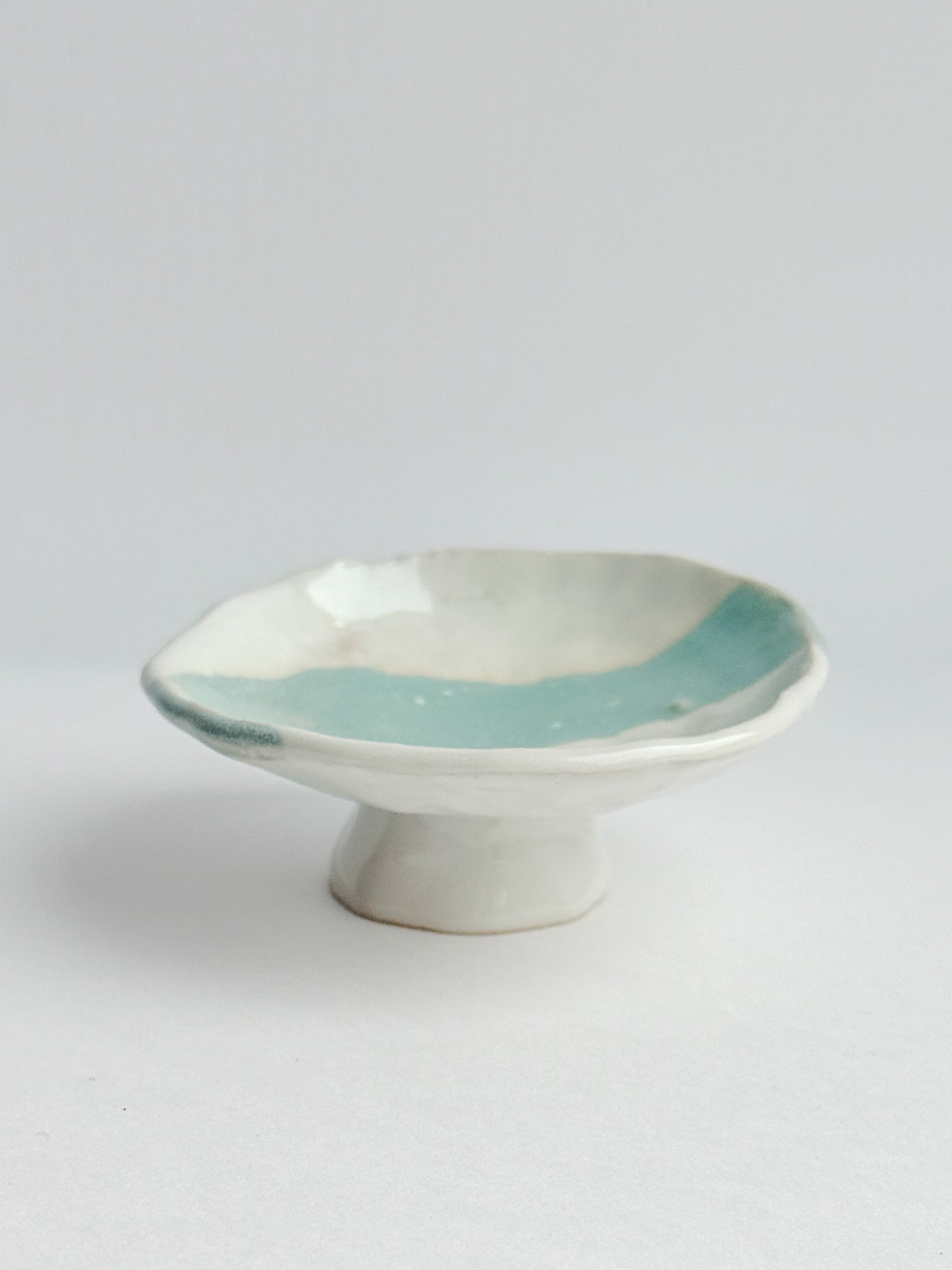 A handmade ceramic pedestal dish with a scalloped rim, featuring a glossy light blue glazed interior and a matte off-white exterior, set against a plain light gray background.