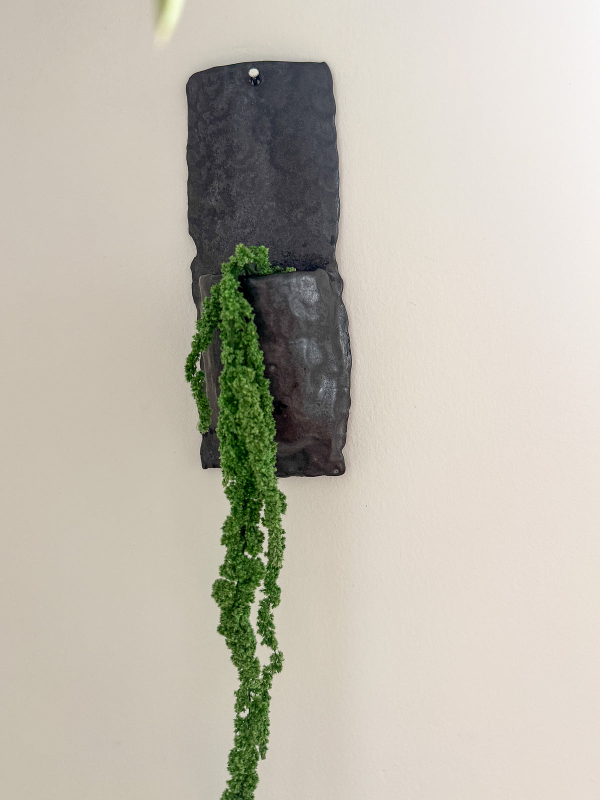 A dark charcoal gray stone wall-mounted planter with a textured, rustic finish, holding a trailing green succulent plant against a plain off-white wall.