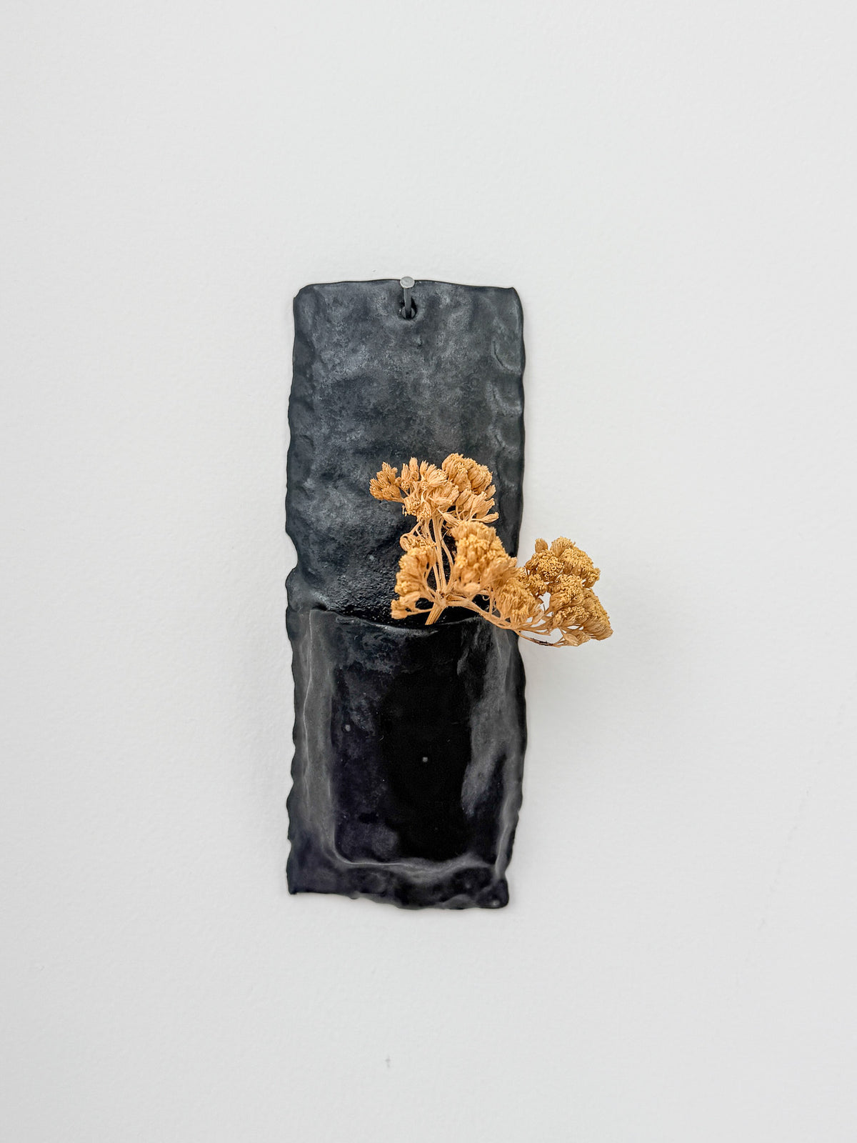 A dark charcoal gray textured ceramic wall-mounted planter with a small dried orange-brown plant inserted, shown against a plain white background.