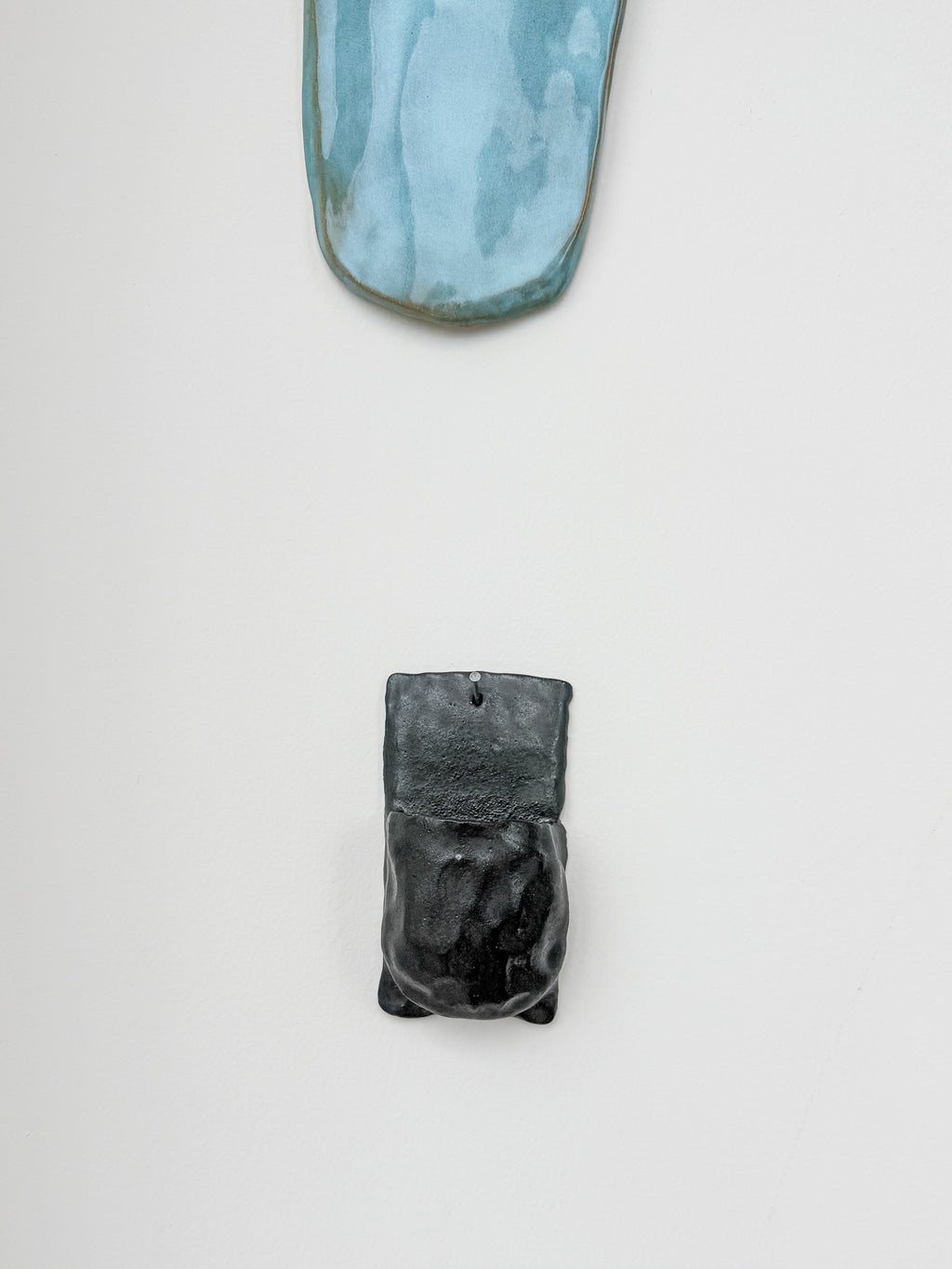 Two ceramic wall-mounted art pieces displayed on a clean white wall. The top piece is a curved, elongated shape with a light blue and gray marbled glaze.
