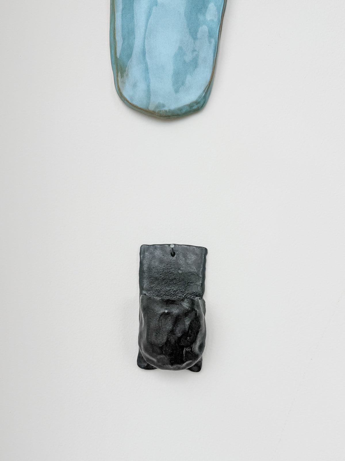 Two ceramic wall-mounted art pieces displayed on a clean white wall. The top piece is a curved, elongated shape with a light blue and gray marbled glaze.