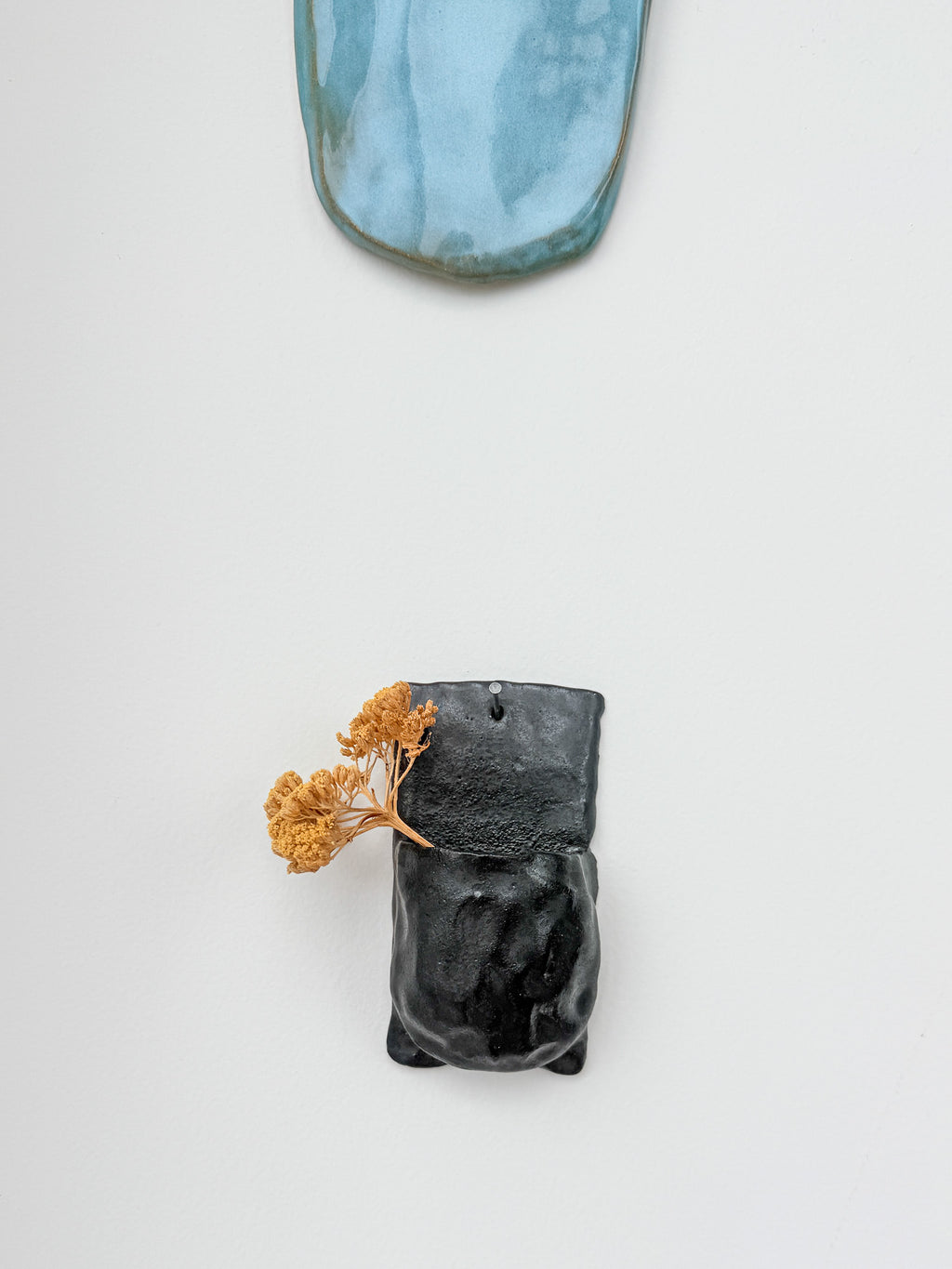 A black ceramic wall-mounted planter with a textured, organic shape, holding dried orange flowers, against a clean white background with a blue-green ceramic tile partially visible at the top.