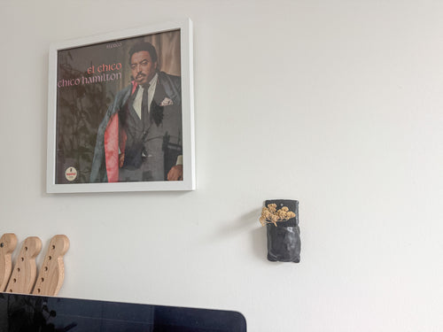 A framed square wall art print featuring a portrait of Chico Hamilton on a record sleeve, hung on a white wall. Part of a wooden headstock and a dark monitor are visible at the bottom, and a small black wall-mounted planter with dried flowers is to the right.