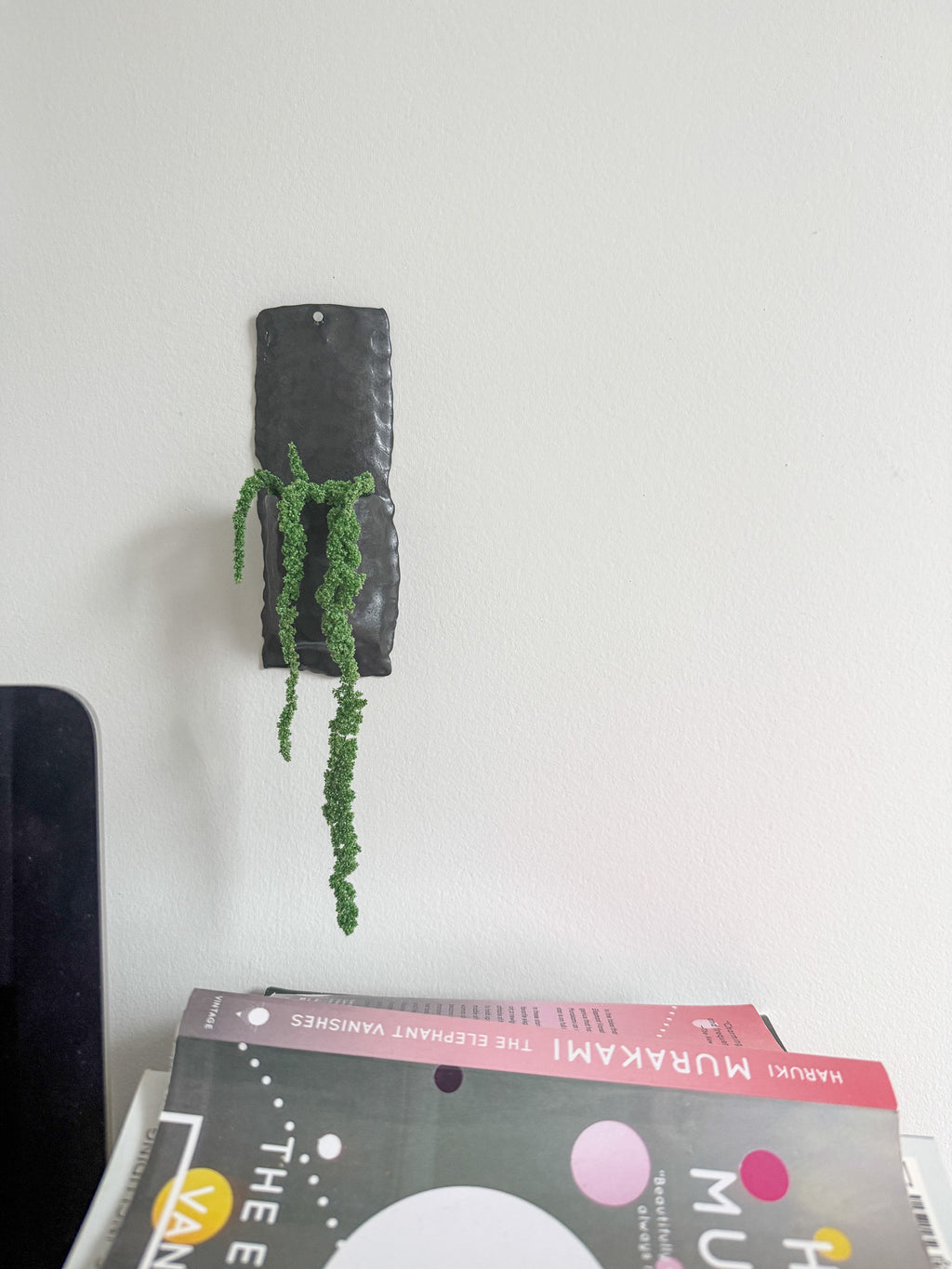 A small black ceramic wall hook shaped like a pillow, holding a dried beige plant, mounted on a plain white wall.