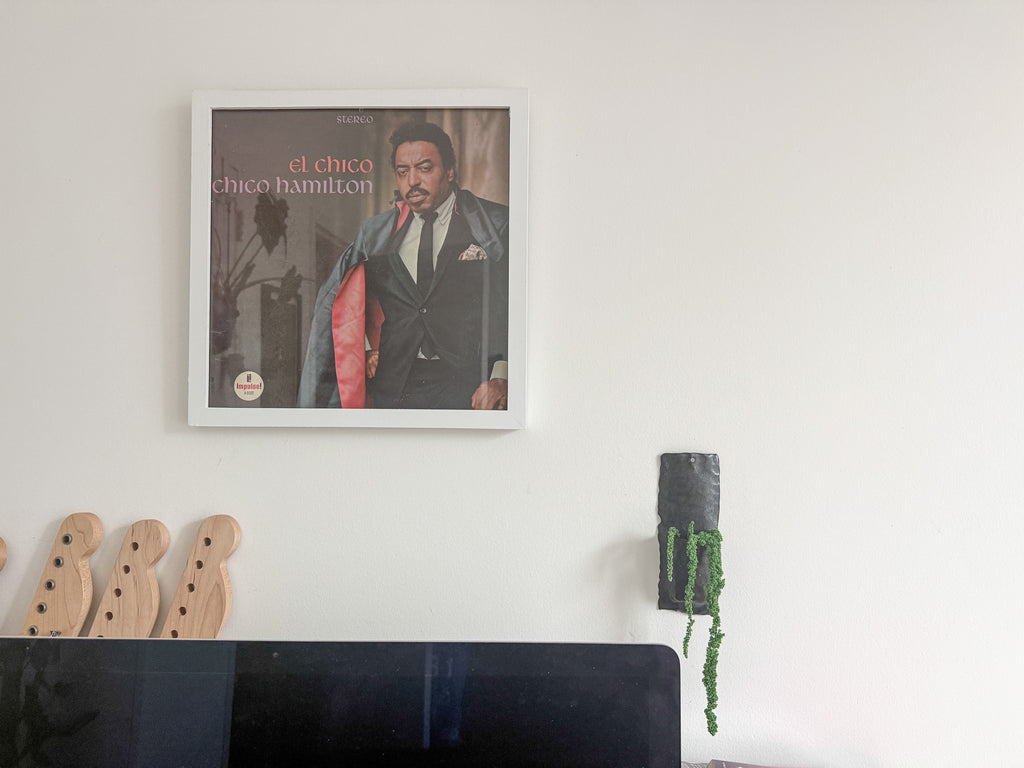 A framed square wall art print featuring a portrait of Chico Hamilton with the text 'el chico chico hamilton' on the cover, hung on a white wall above a dark surface. Part of a wooden object and a hanging green plant are visible at the bottom.