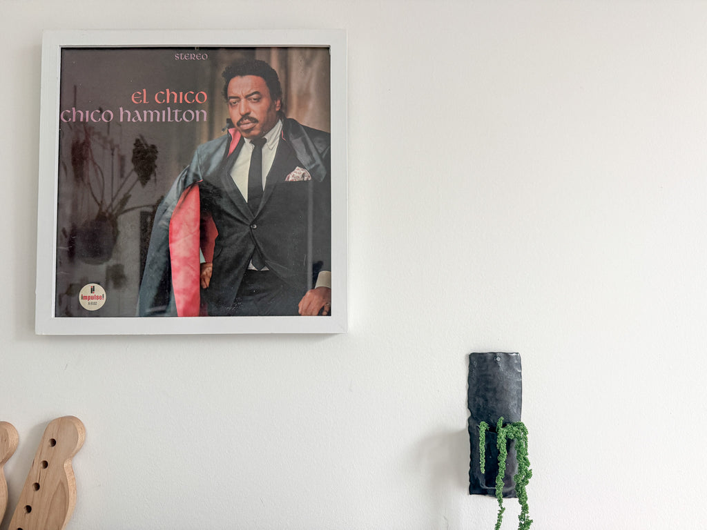 A framed album cover of Chico Hamilton's 'El Chico' hanging on a white wall, with part of a wooden headstock and a small green hanging plant visible in the room.
