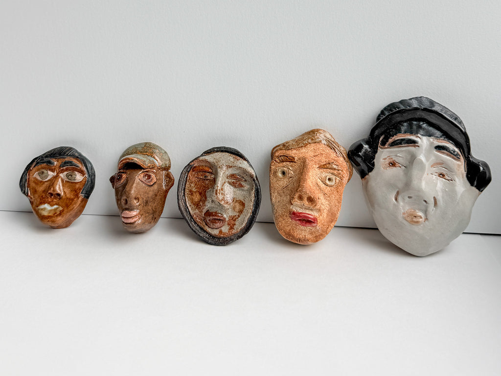 Five handcrafted ceramic masks with stylized human faces, arranged in a row against a plain white background.