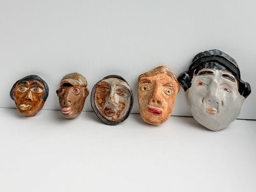 Five handcrafted ceramic masks with stylized human faces, arranged in a row against a plain white background.