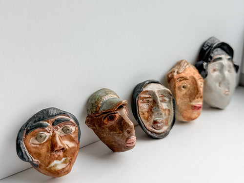 Five small sculptural ceramic faces in varied earthy tones are lined up against a plain white wall and surface.