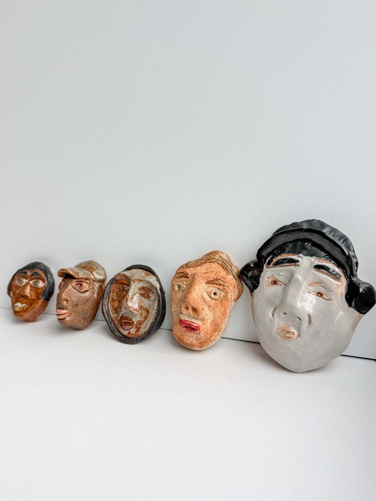 Five handcrafted ceramic masks with expressive faces are lined up in a row against a plain light gray background. The masks have varied earthy tones including brown, beige, and gray, with one mask on the right wearing a black cap.