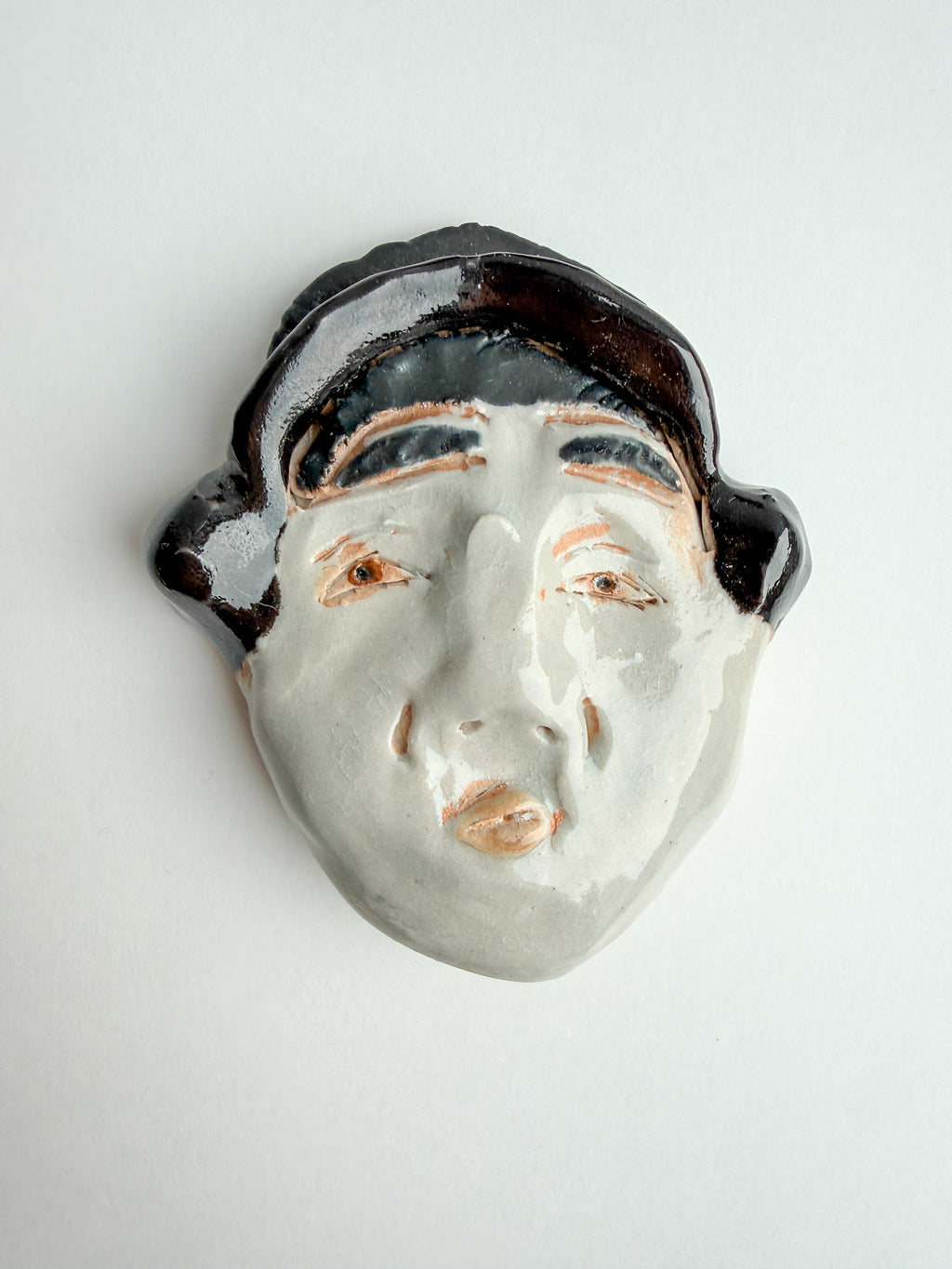 A ceramic face wall sculpture with a glossy white face, dark brown hair, and peach-colored eyes and lips, shown against a plain light gray background.