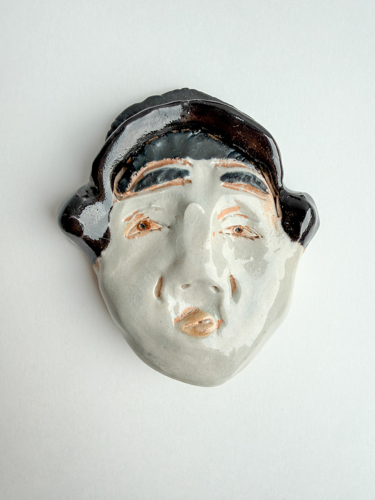 A ceramic face wall sculpture with a glossy white face, dark brown hair, and peach-colored eyes and lips, shown against a plain light gray background.
