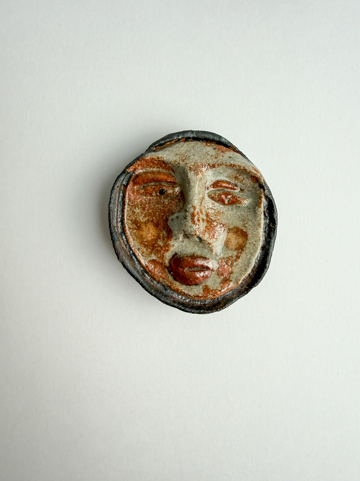 A round, handcrafted ceramic wall plaque featuring a stylized face with a textured, weathered finish in earthy brown, beige, and black tones, set against a plain off-white background.