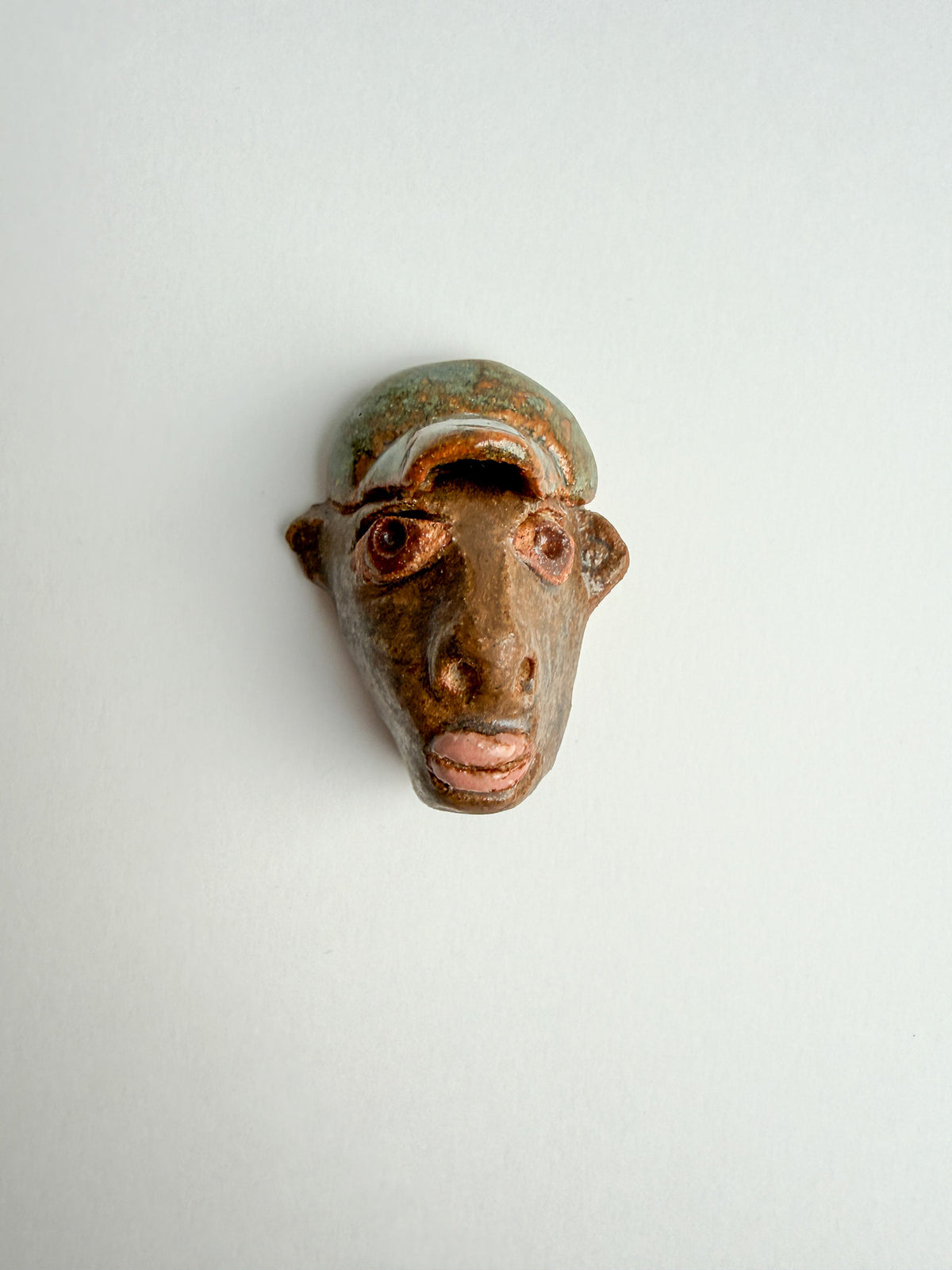 A small handmade ceramic face mask or sculptural head with a glossy finish, featuring brown and green tones, photographed against a plain off-white background.