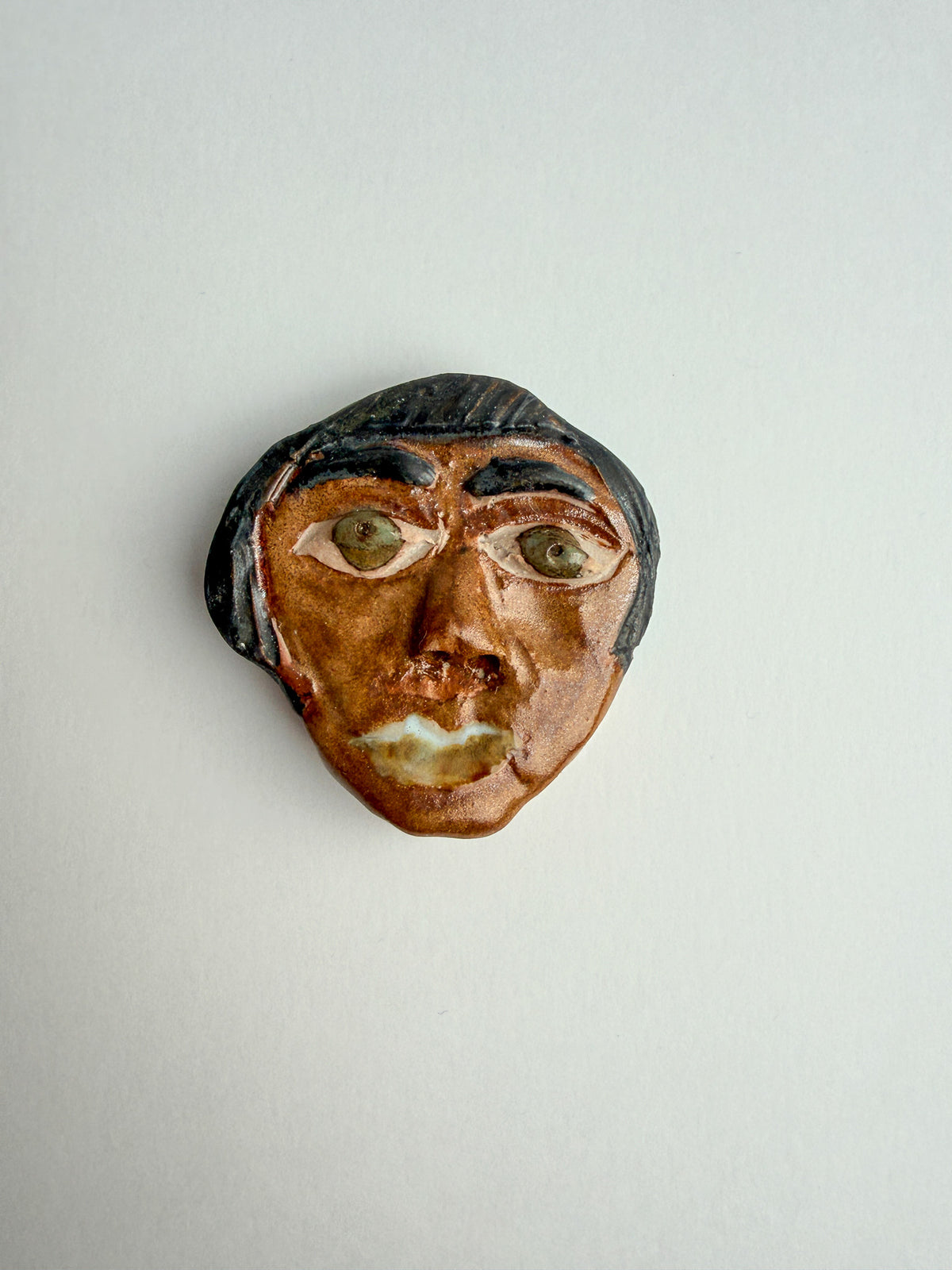 A small sculptural ceramic face with a brown skin tone, black hair, and green eyes, photographed against a plain light gray background.