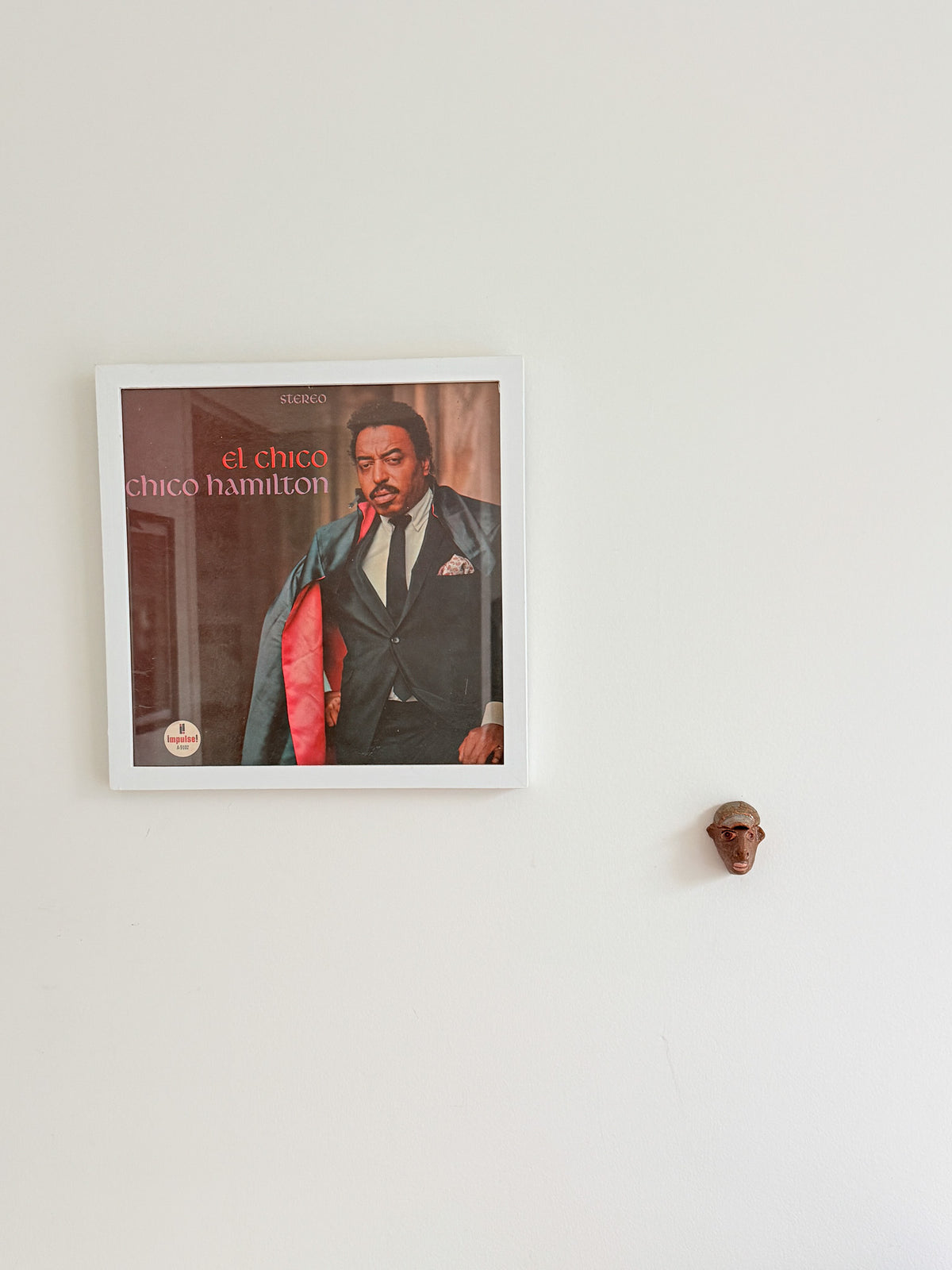 A framed square wall art print featuring a portrait of Chico Hamilton on an album cover, hung on a plain white wall. A small brown sculptural object is visible to the right of the frame.