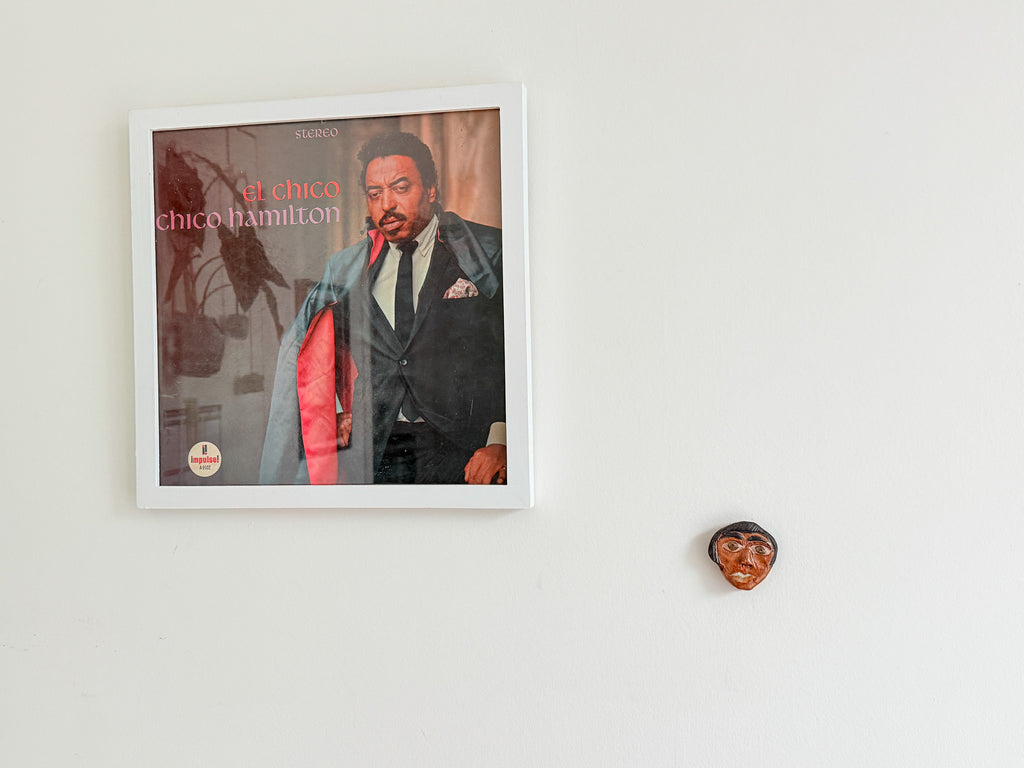 A framed album cover featuring Chico Hamilton hangs on a white wall, with a small carved wooden face sculpture to the right.