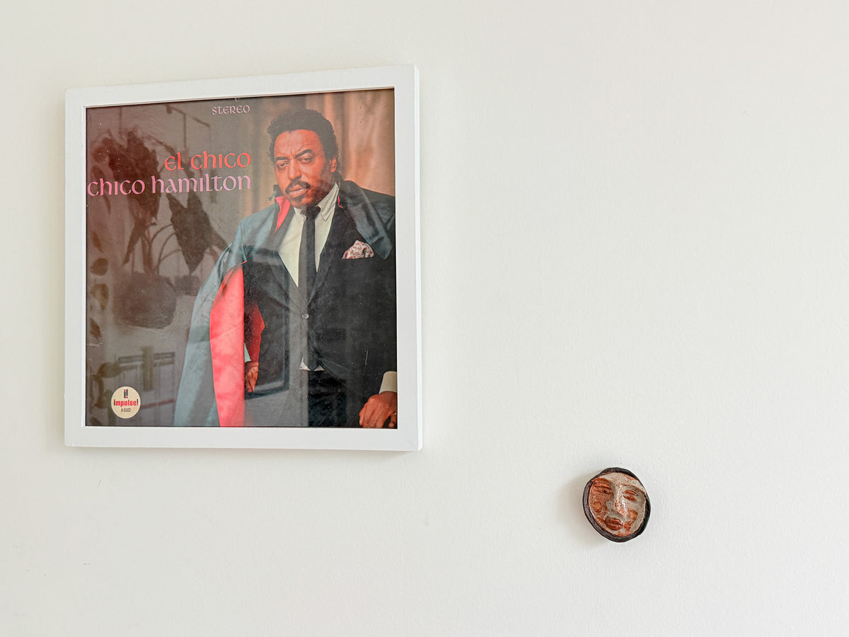A framed square wall art print featuring a portrait of Chico Hamilton, hung on a plain white wall alongside a small round decorative object.