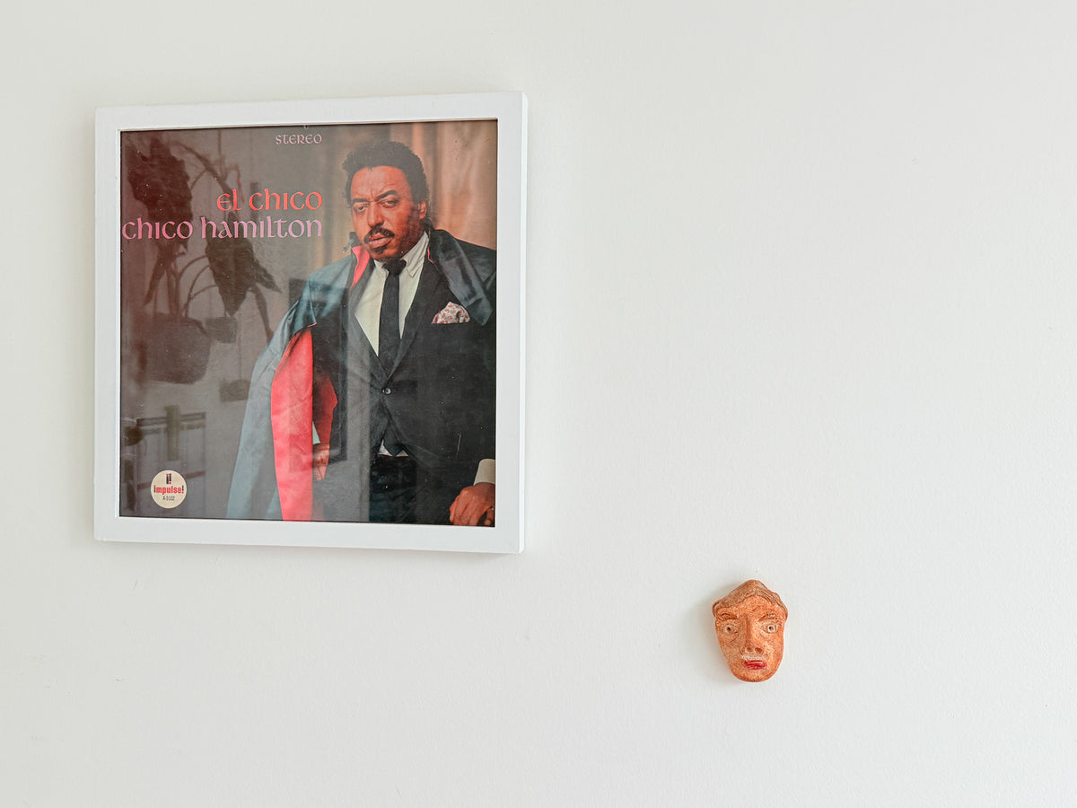 A framed square wall art print featuring a portrait of Chico Hamilton with the text 'el chica chico hamilton' on the cover, mounted on a plain white wall. A small terracotta face sculpture is visible below the frame.