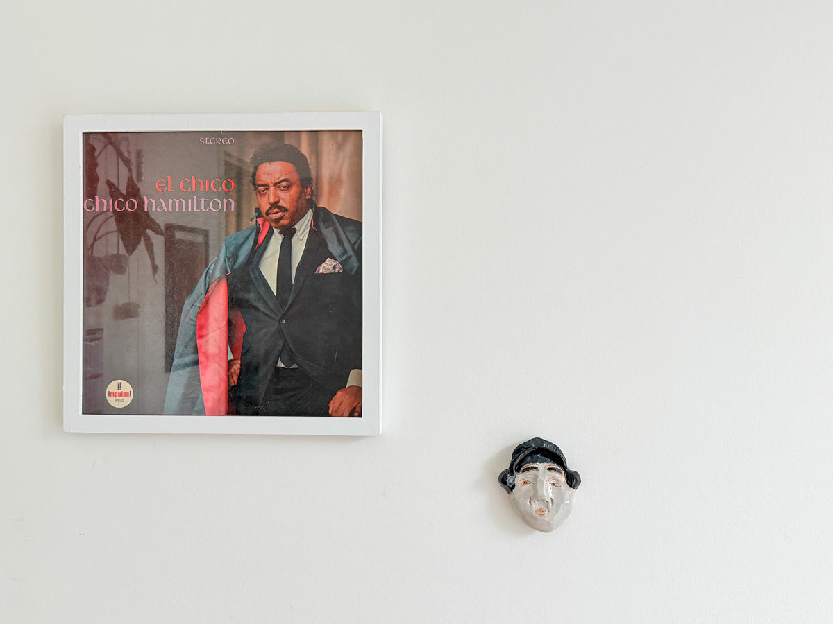 A framed square wall art print featuring a portrait of Chico Hamilton with the text 'el chico chico hamilton' on the cover, mounted on a plain white wall. A small decorative face mask hangs to the right of the frame.