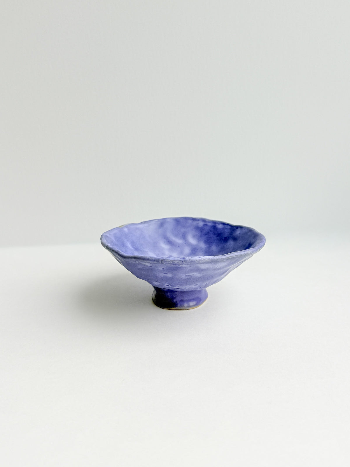 A small handmade ceramic bowl with a textured, mottled blue and purple glaze, standing on a short footed base against a clean white background.