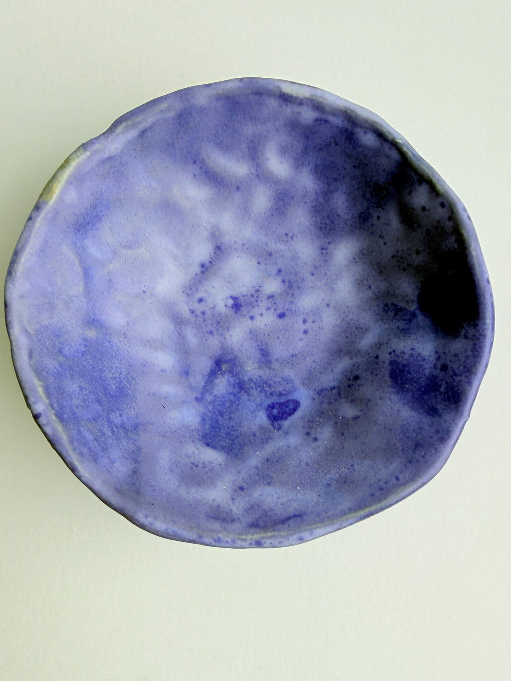 A handmade ceramic bowl with a textured, organic shape, featuring a glaze of mottled light blue, lavender, and deep purple with dark blue speckles, shown from a top-down view on a light beige background.