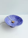 A silver chain necklace with a small, textured gold pendant displayed on a blue ceramic bowl against a light gray background.