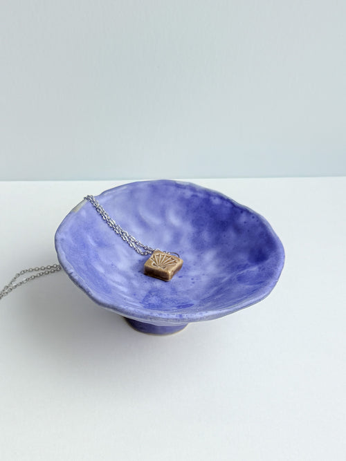 A silver chain necklace with a small, textured gold pendant displayed on a blue ceramic bowl against a light gray background.