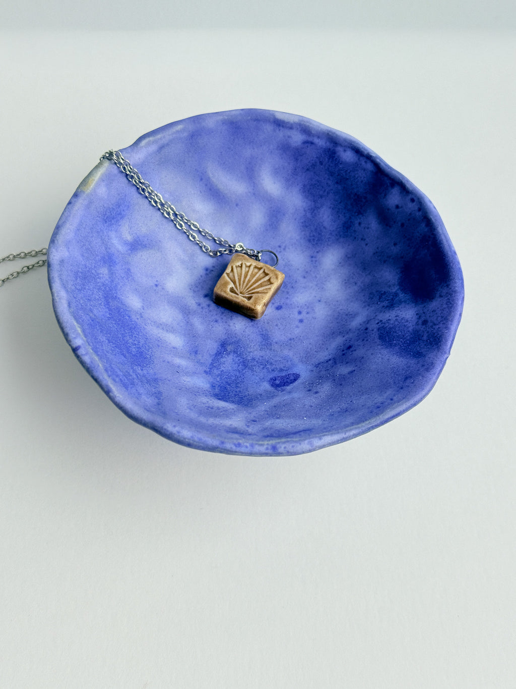 A small pendant necklace with a gold-toned fan-shaped charm on a silver chain, displayed inside a textured blue ceramic bowl against a plain white background.